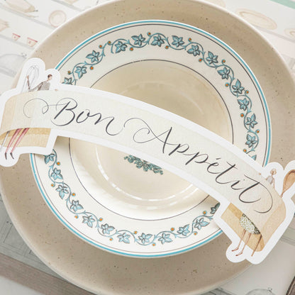 Two Cooks Banner Table Accent with &quot;Bon Appetit&quot; written on it, resting on dinner plates, on an elegant place setting for one.