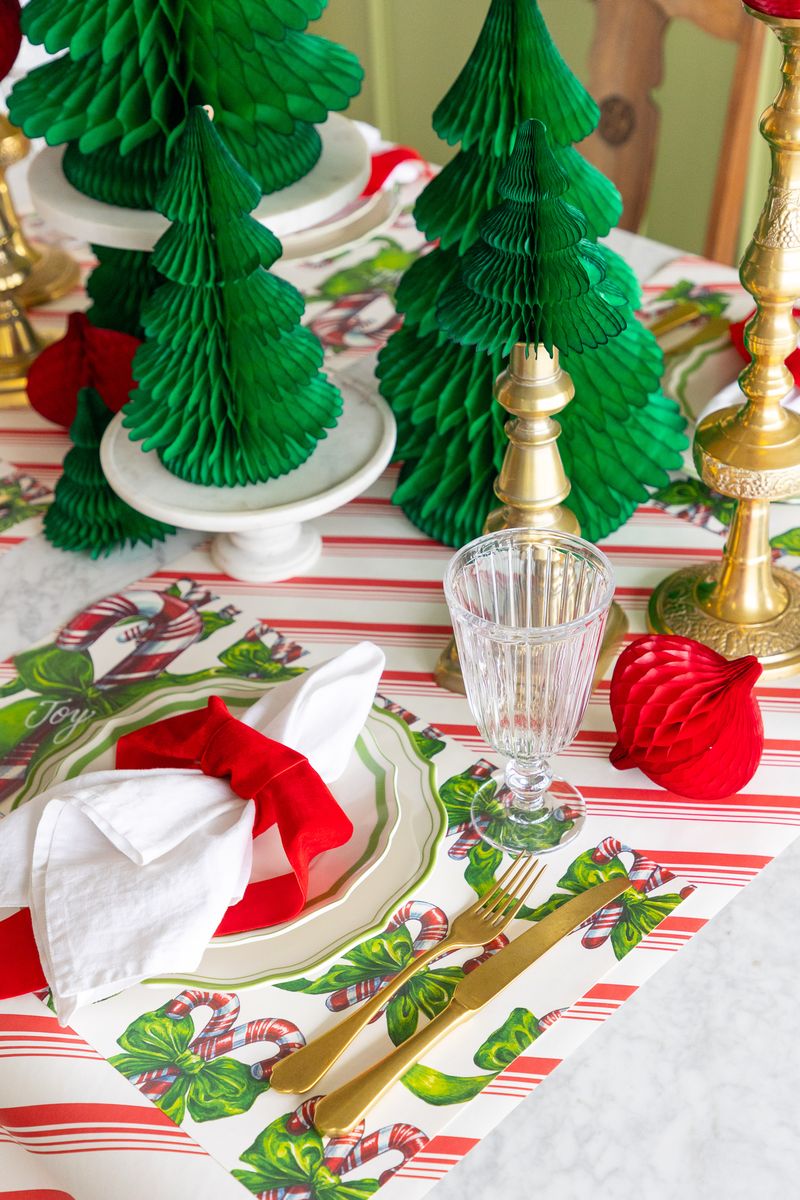 Christmas-themed table setting with green paper trees, gold cutlery, and red ornaments on a patterned tablecloth.