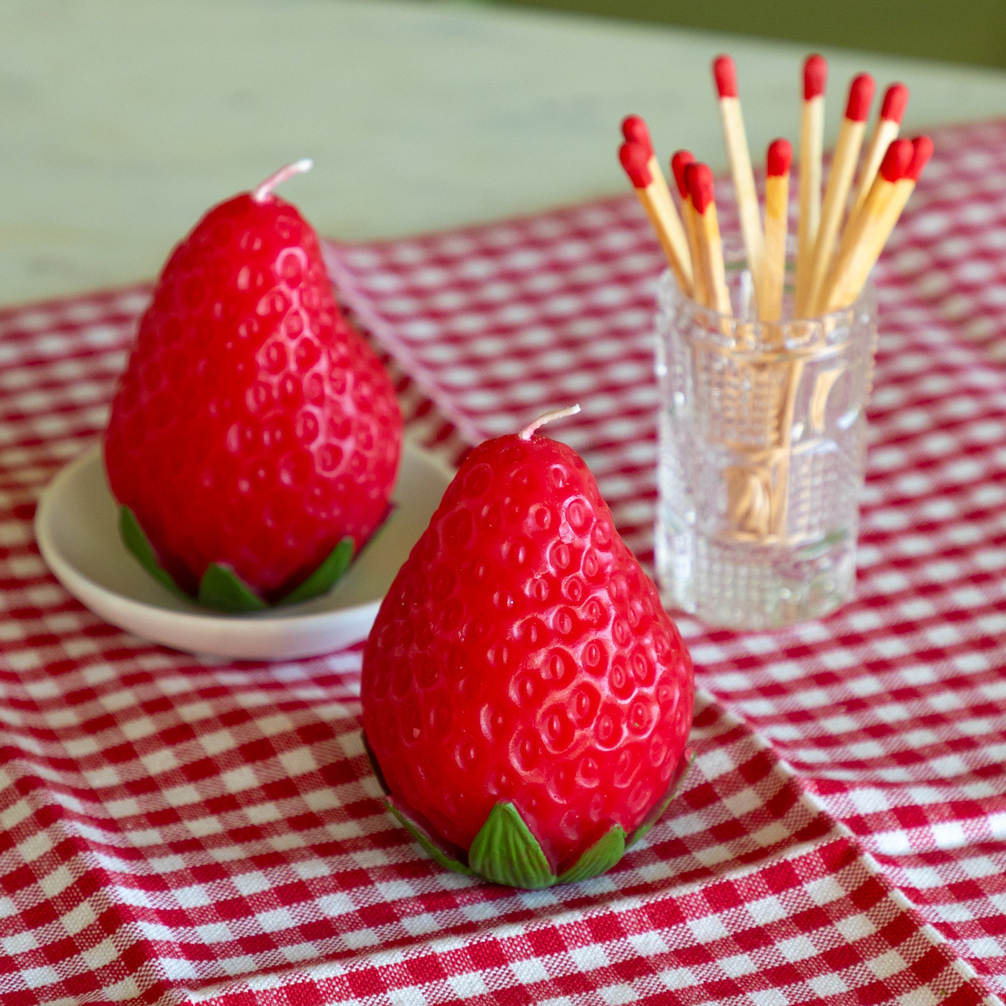 Red strawberry-shaped candles on a red and white checkered tablecloth with a small glass container of matches.