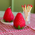 Two strawberry-shaped candles and a cup of matches on a table with a red and white checkered tablecloth.