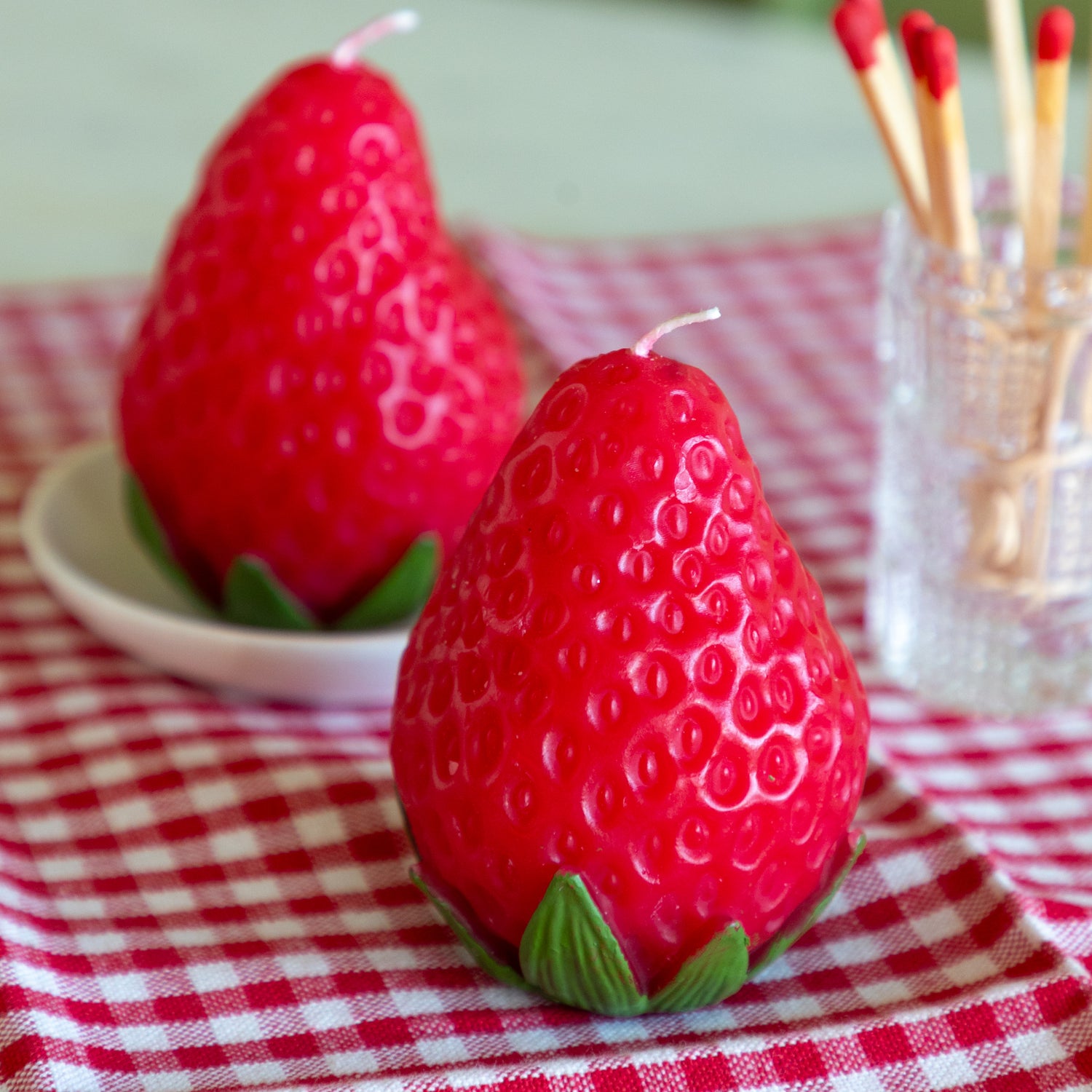 Strawberry-shaped candles on a red and white checkered tablecloth with matches in the background.