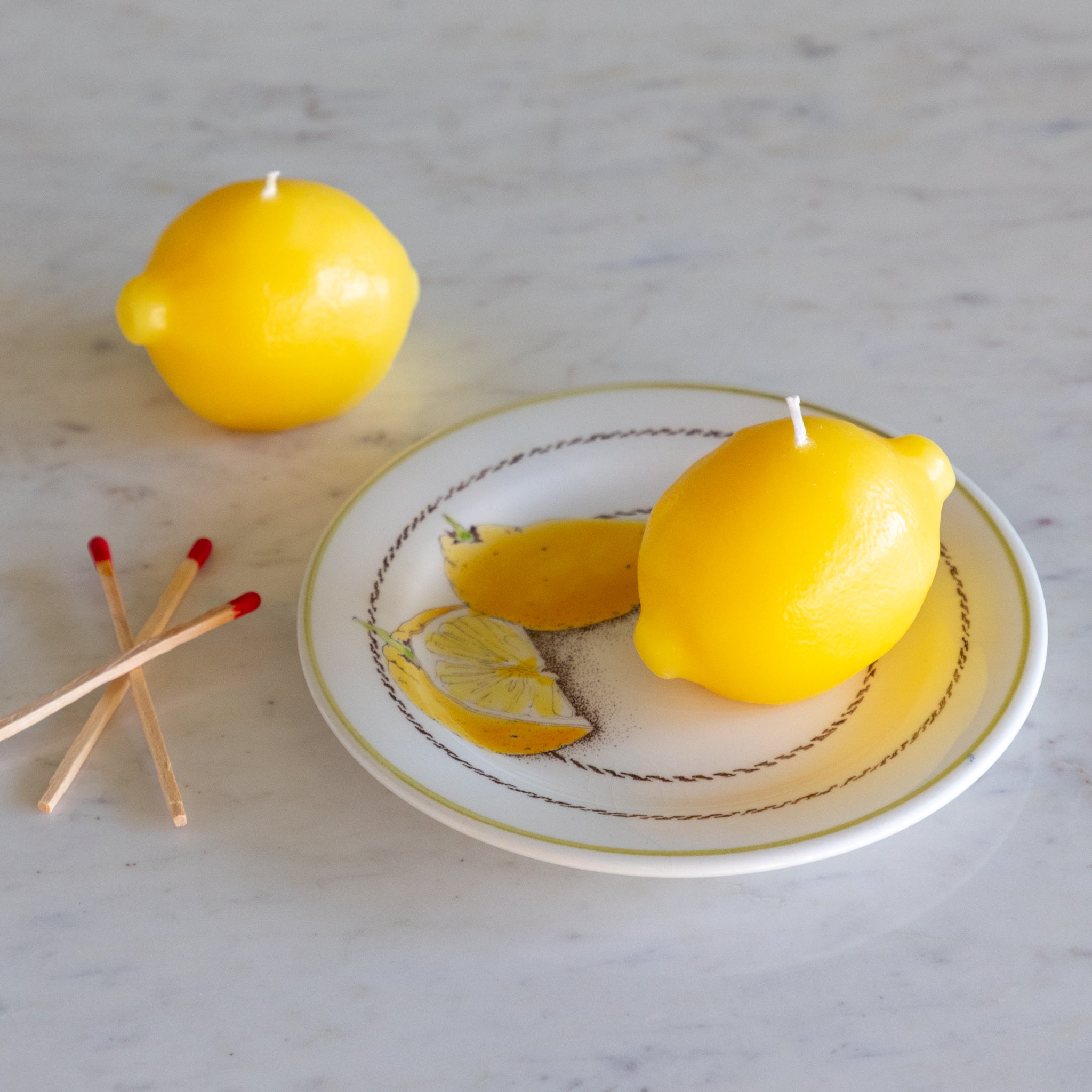Two lemon-shaped candles on a decorative plate with matches on a marble surface.