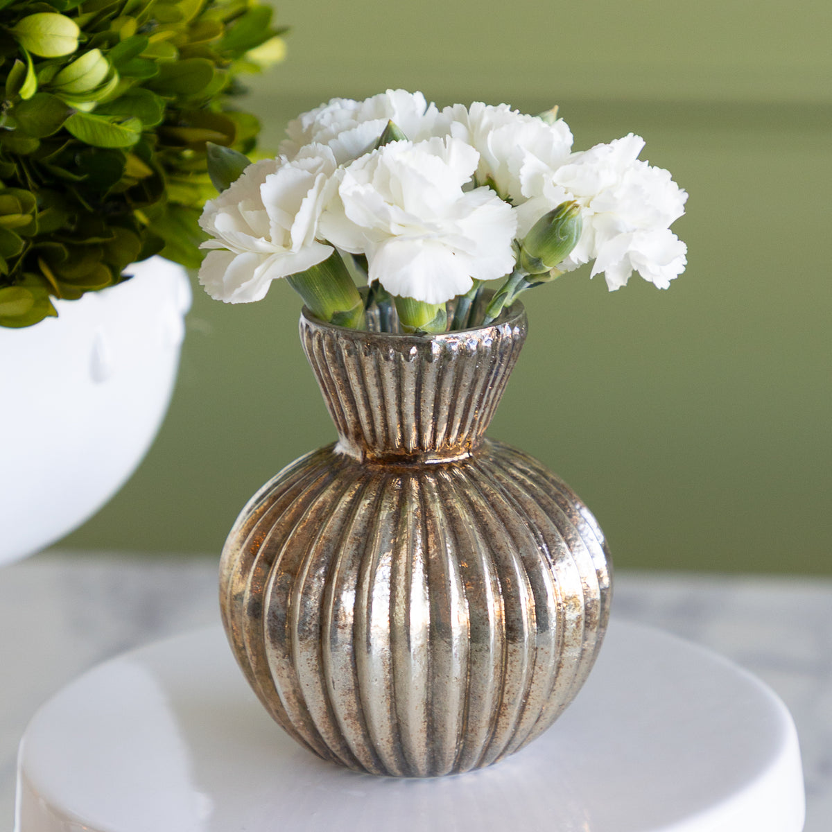 A ribbed metallic gold ceramic vase filled with white carnation flowers, displayed on a round white pedestal against a soft green background.
