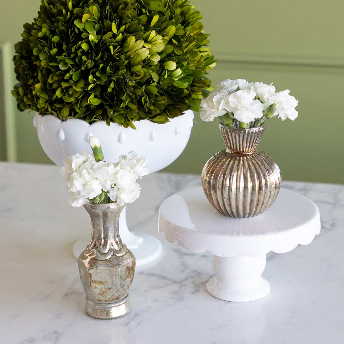 Three decorative vases and floral arrangements displayed on a white marble surface against a sage green wall. The arrangement includes a large round green boxwood top in a white pedestal bowl, white carnations in a ribbed metallic gold vase on a white cake stand, and white carnations in a small distressed gold vase.