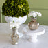 Three decorative vases and floral arrangements displayed on a white marble surface against a sage green wall. The arrangement includes a large round green boxwood top in a white pedestal bowl, white carnations in a ribbed metallic gold vase on a white cake stand, and white carnations in a small distressed gold vase.