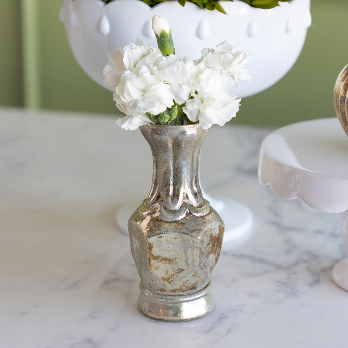 A small distressed silver metal vase with white carnations on a white marble surface, with a white ceramic bowl in the background.
