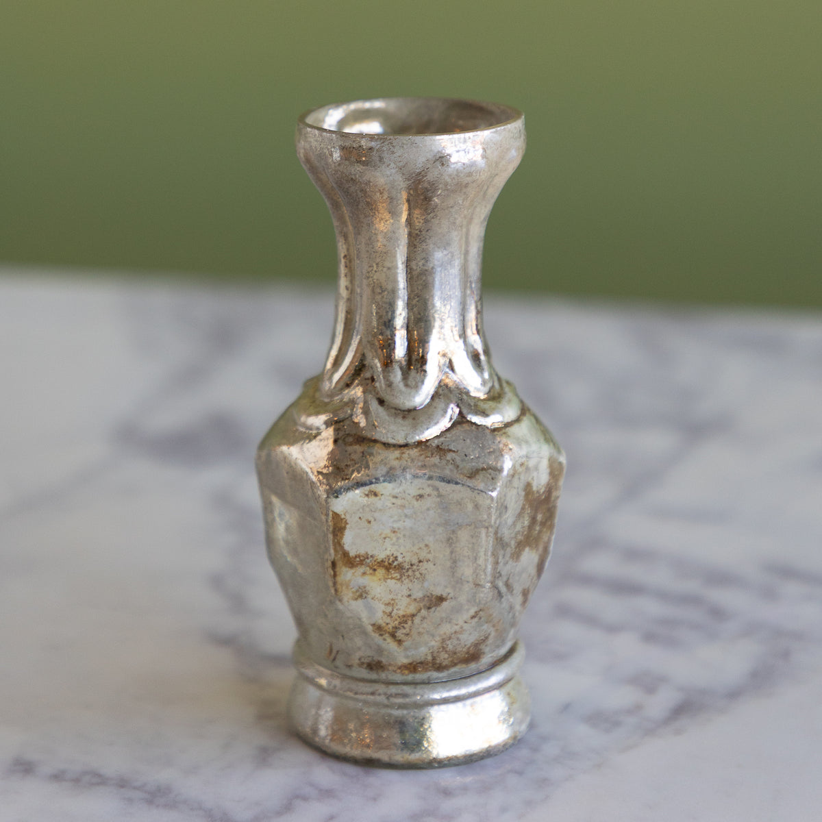 A small vintage-style metallic vase with a distressed silver finish, featuring a flared neck and a textured, weathered body, set on a white marble surface with a soft olive green background.