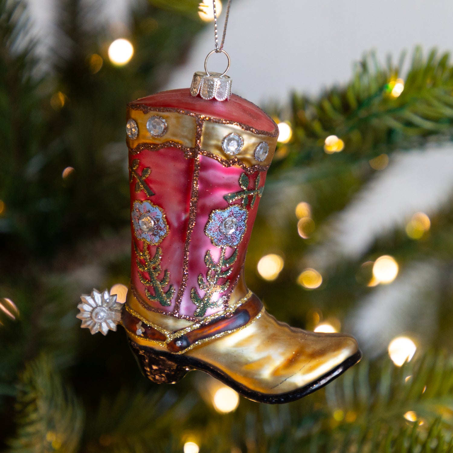 Decorative cowboy boot ornament on a Christmas tree with lights in the background.