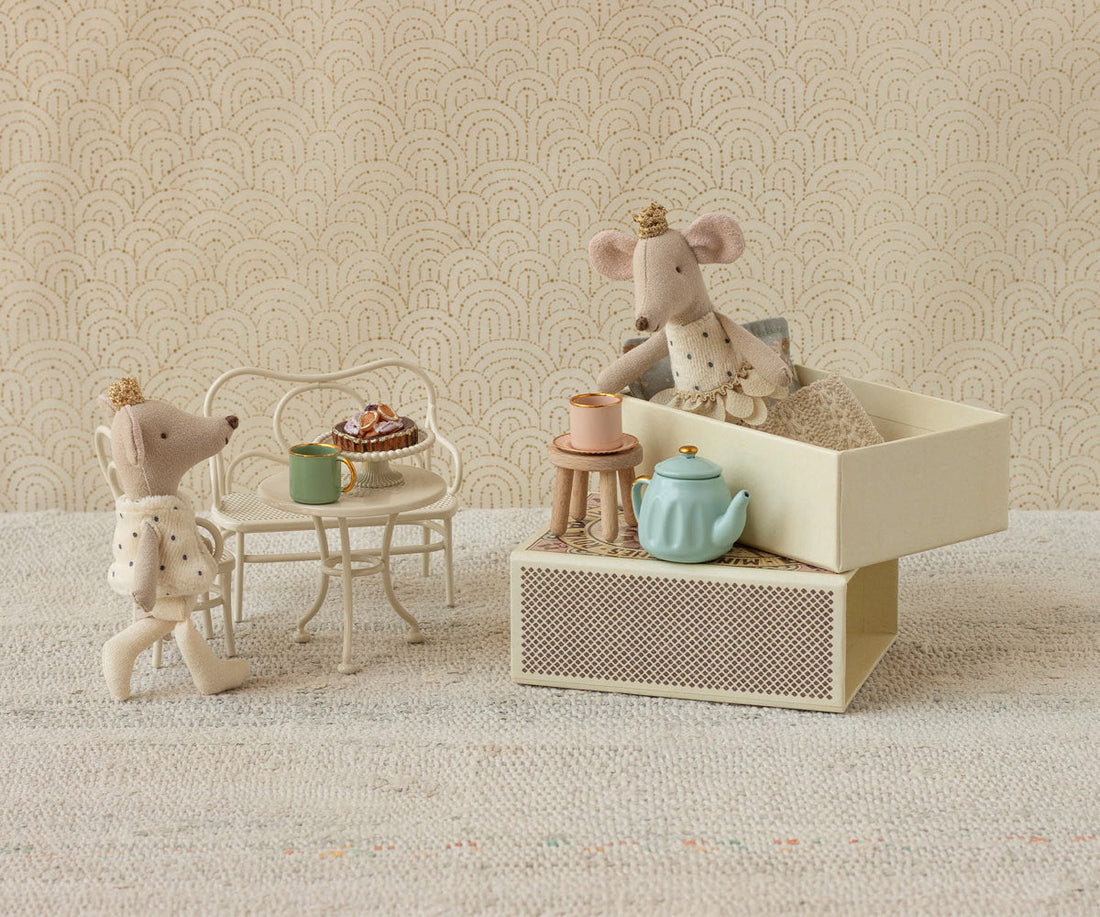 Two toy mice with a miniature tea set on a textured surface with a patterned wall background.