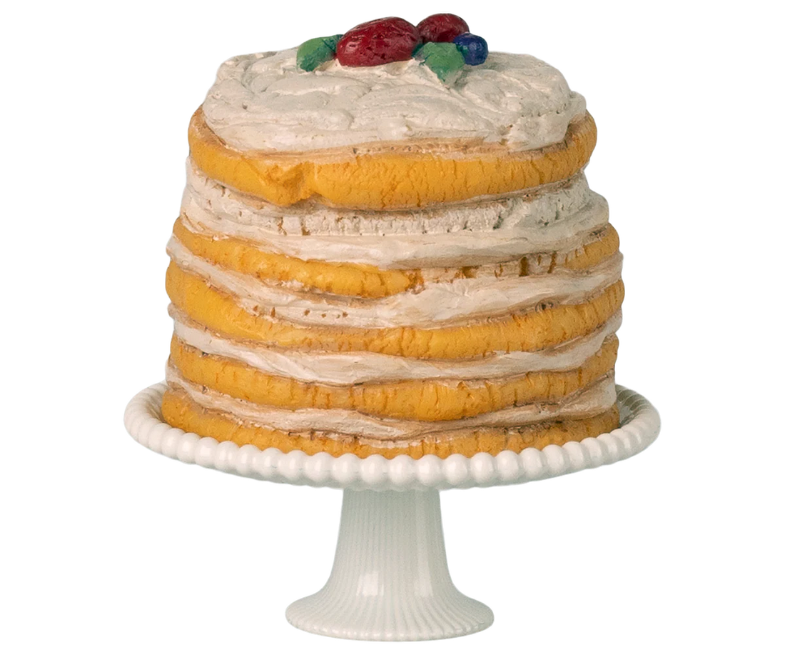A miniature toy layered cake with white frosting and fruit on top, placed on a white cake stand.