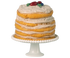A miniature toy layered cake with white frosting and fruit on top, placed on a white cake stand.