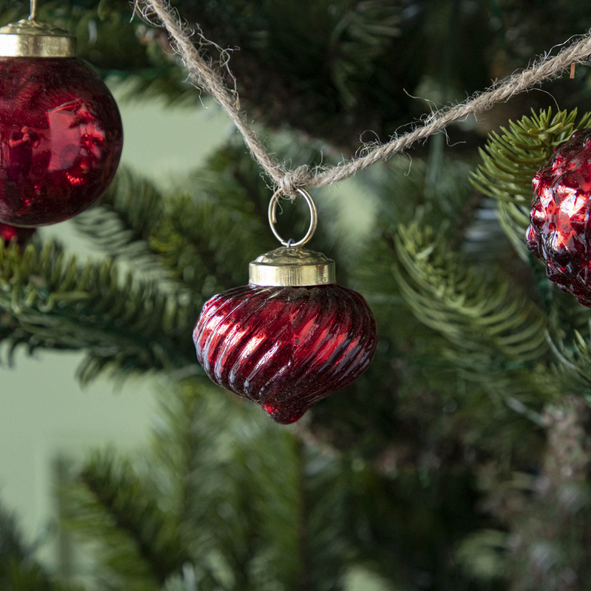 A close up of three Embossed Red Mercury Glass Ornaments showing the detailing and jute cord hanging on a tree.