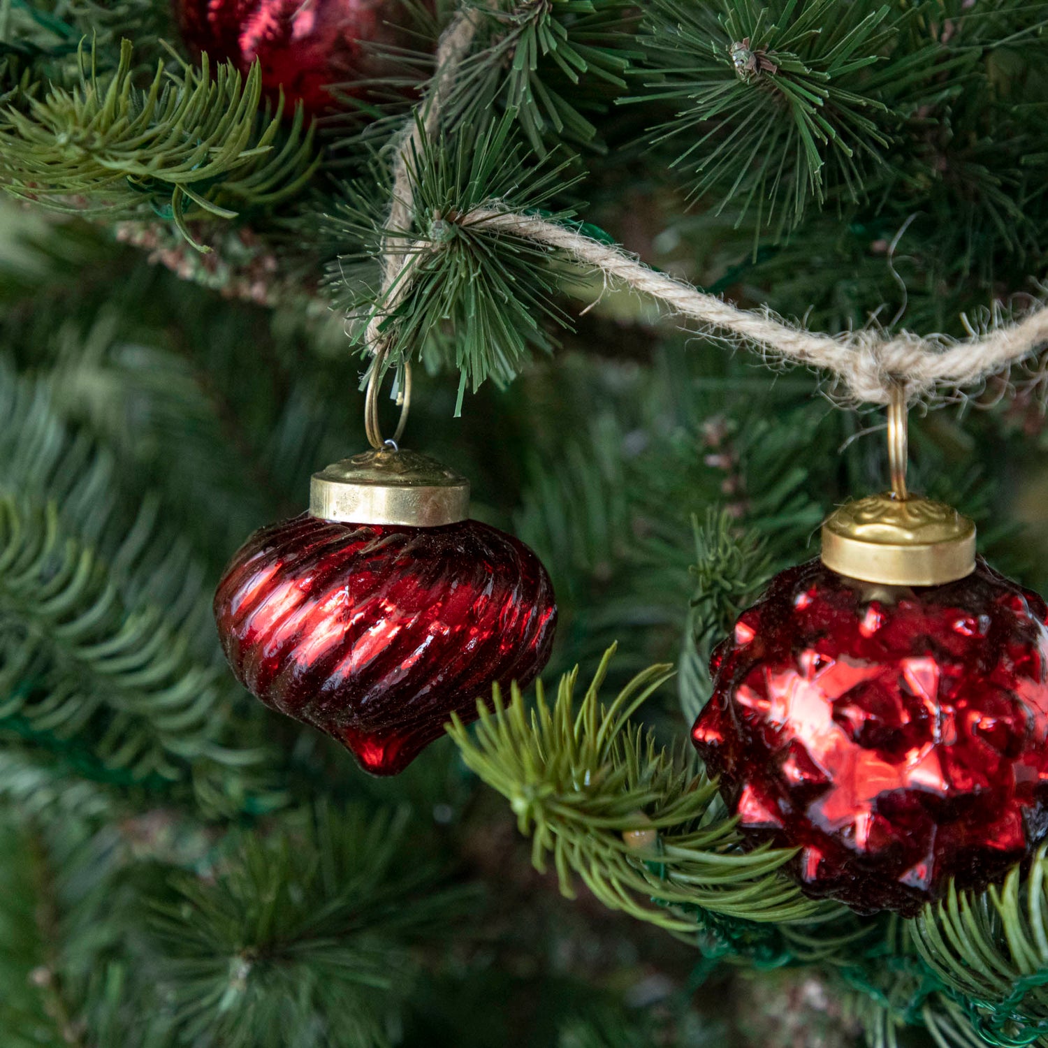 A close up of two Embossed Red Mercury Glass Ornaments showing the ribbed and spiked detailing and jute cord hanging on a tree.