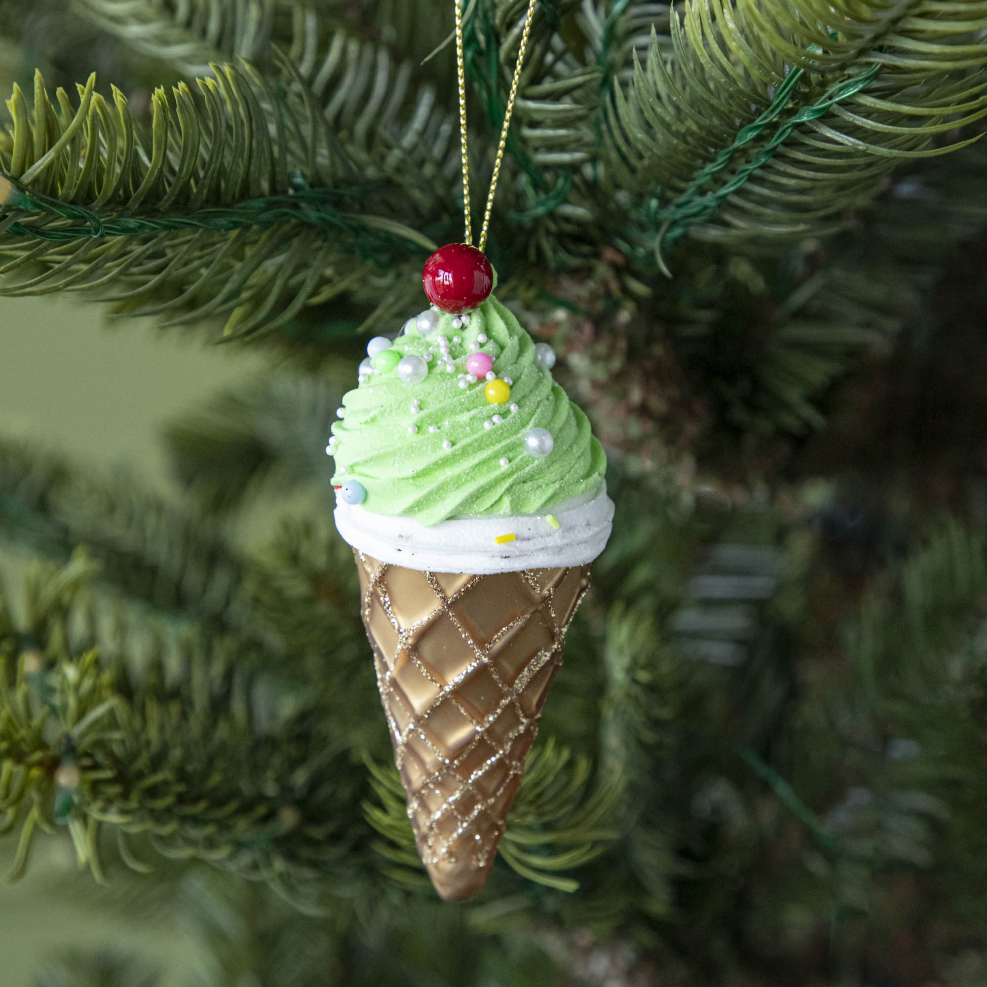 An Ice Cream Cone ornament with a sparkled brown cone, green and white icing and sprinkles with a cherry on top, hanging on a tree by a green string.