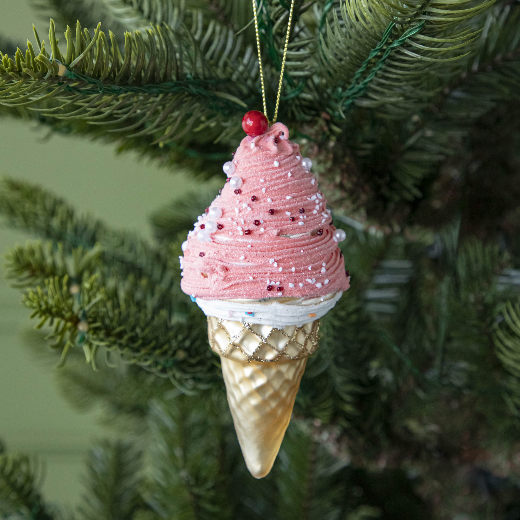 An Ice Cream Cone ornament with a sparkled tan cone, pink and white white icing with sprinkles and a cherry on top, hanging on a tree by a green string.