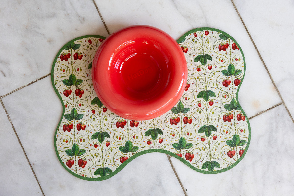 A red ceramic bowl placed on a decorative bone-shaped placemat with a strawberry and leaf pattern, set on a light marble tile floor.