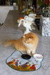 A fluffy orange cat stands on a white marble kitchen counter next to a cat-shaped cat mat with colorful bird illustrations, a black bowl, and a white dish. A white swan figurine and other kitchen items are blurred in the background.