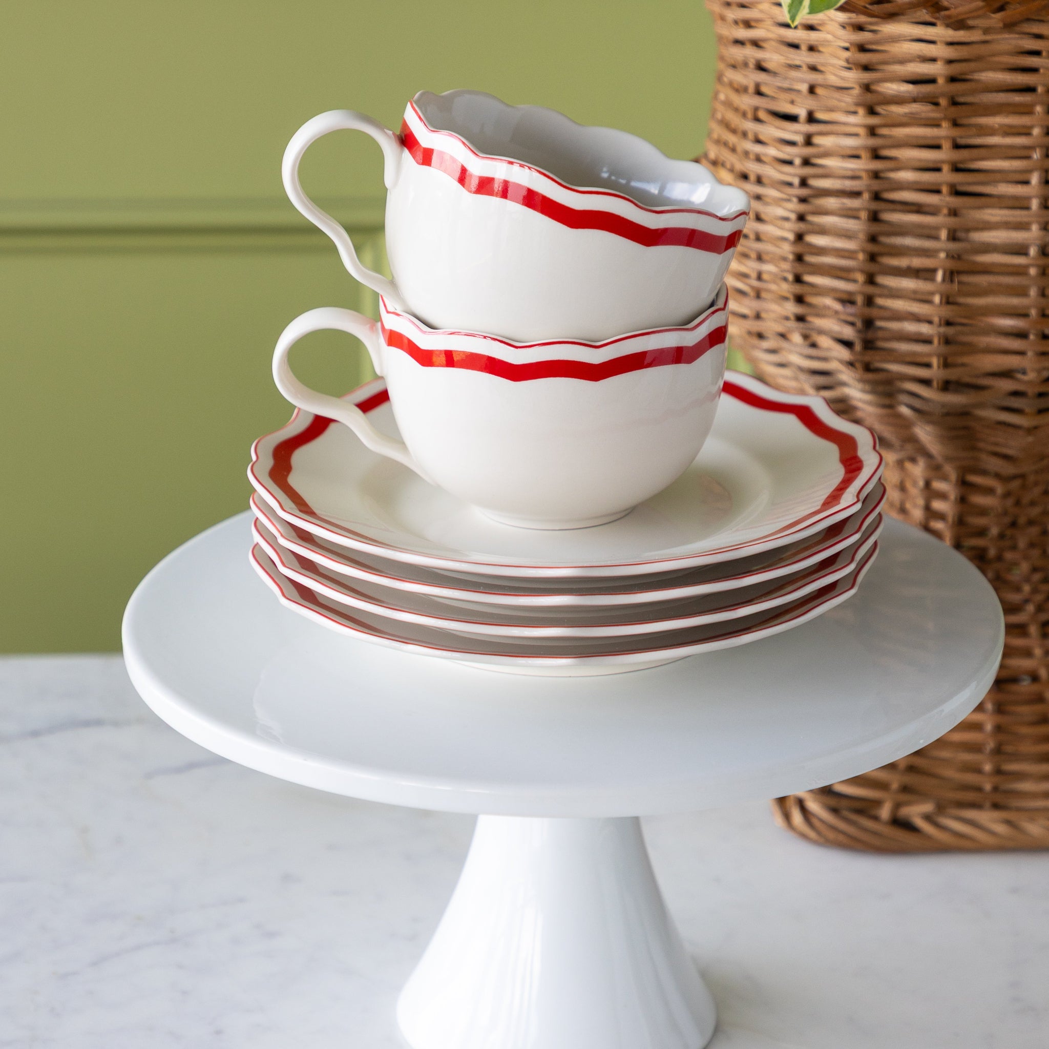 Stack of white ceramic cups and saucers with red stripes on a white cake stand against a green wall.