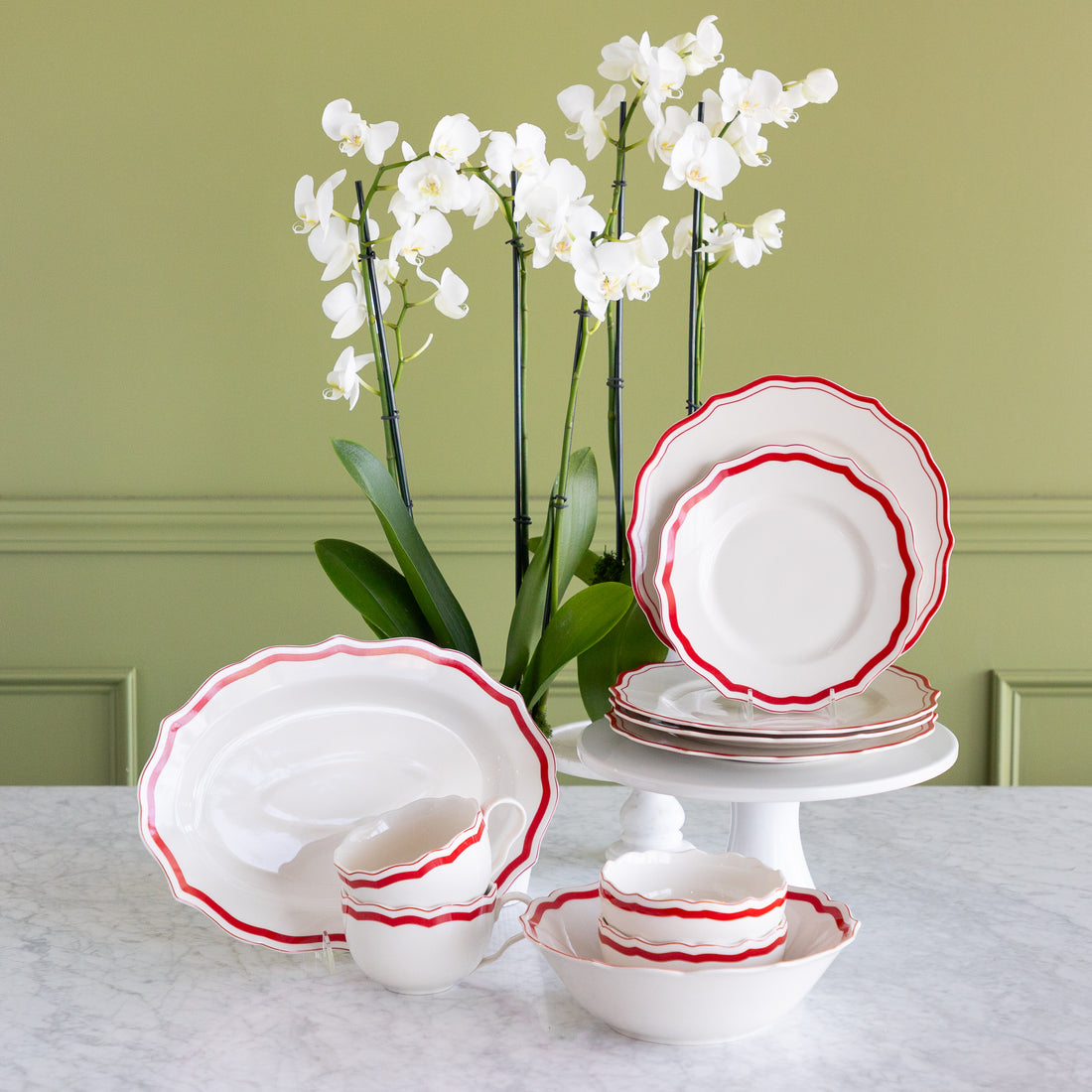 White ceramic dinnerware set with red scalloped edges, including plates, bowls, cups, and a platter, arranged on a marble table with white orchids against a green wall.