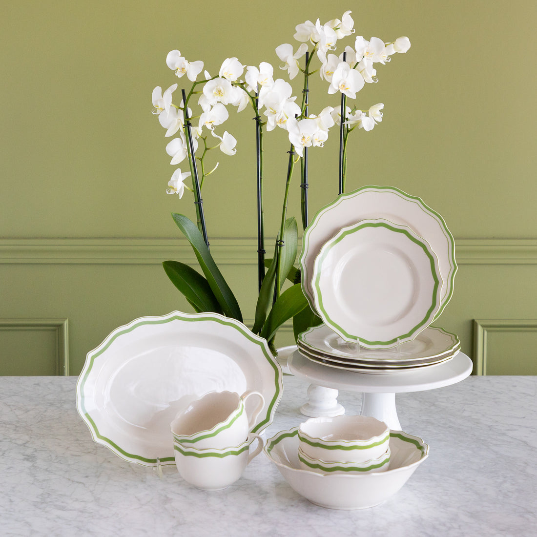 A coordinated white dinnerware set with a green scalloped rim, displayed on a marble table against an olive green paneled wall. The set includes plates, bowls, cups, and a platter, styled with white orchids in the background.
