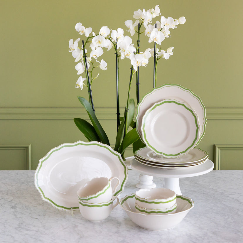 A coordinated white dinnerware set with a green scalloped rim, displayed on a marble table against an olive green paneled wall. The set includes plates, bowls, cups, and a platter, styled with white orchids in the background.