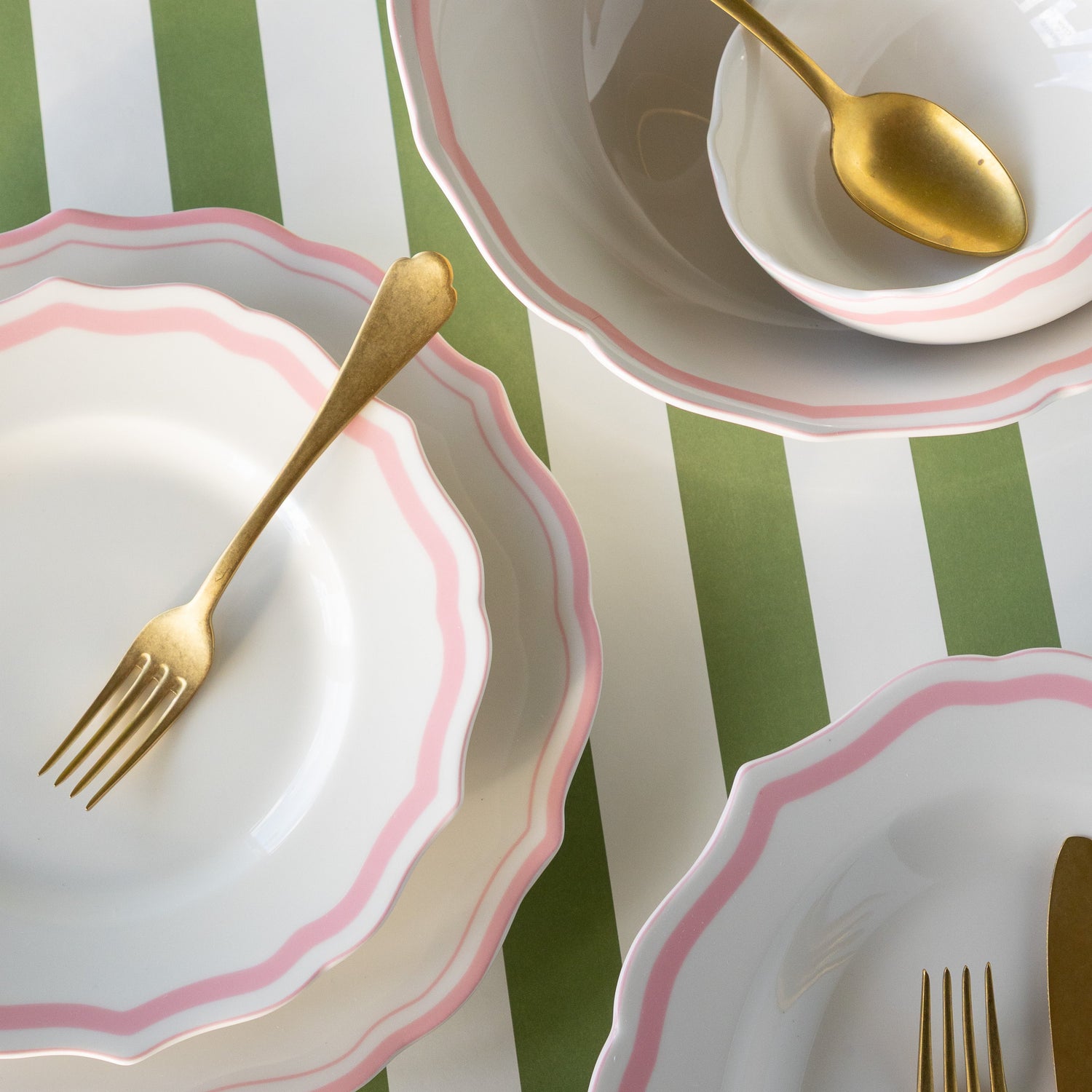 Set of white plates with pink rims and gold cutlery on a green and white striped background.