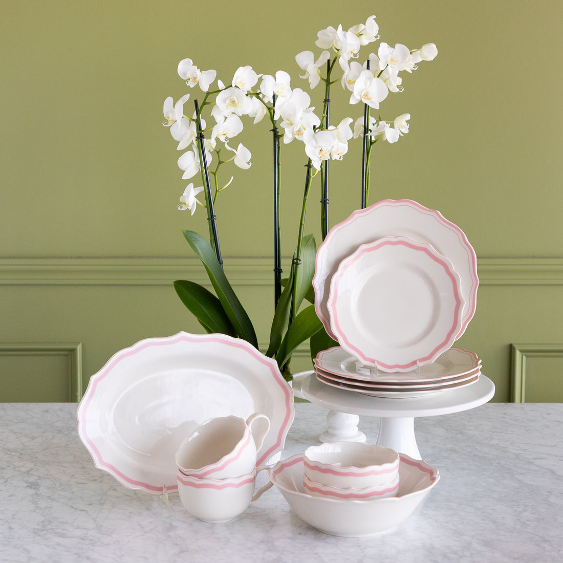 A coordinated white dinnerware set with a delicate pink scalloped rim, displayed on a marble table against an olive green wall. The set includes plates, bowls, cups, and a platter, styled with white orchids in the background.