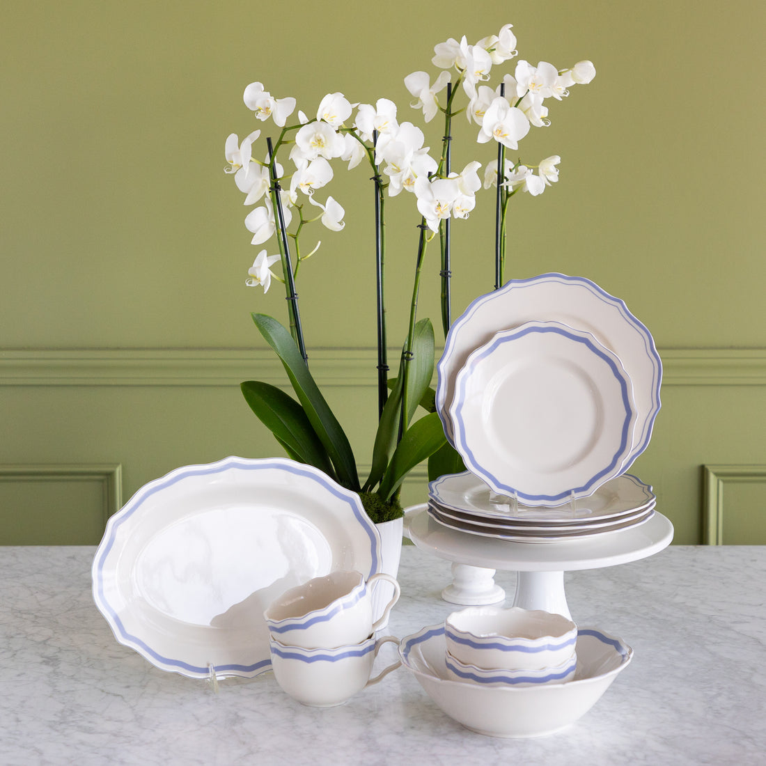 A white porcelain dinnerware set with blue scalloped rims, including plates, bowls, cups, and a platter, arranged on a marble table with white orchids against a sage green paneled wall.