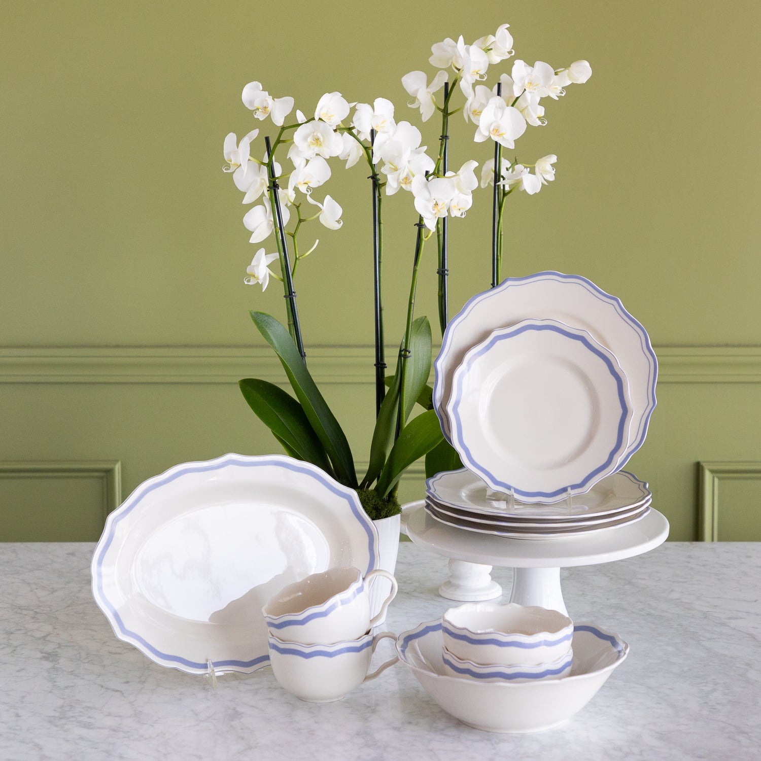 A white porcelain dinnerware set with blue scalloped rims, including plates, bowls, cups, and a platter, arranged on a marble table with white orchids against a sage green paneled wall.