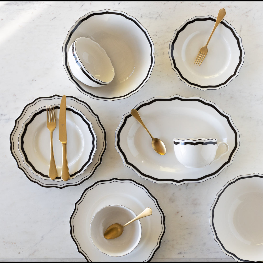 Set of white ceramic dishes with black rims and gold cutlery on a light gray surface.