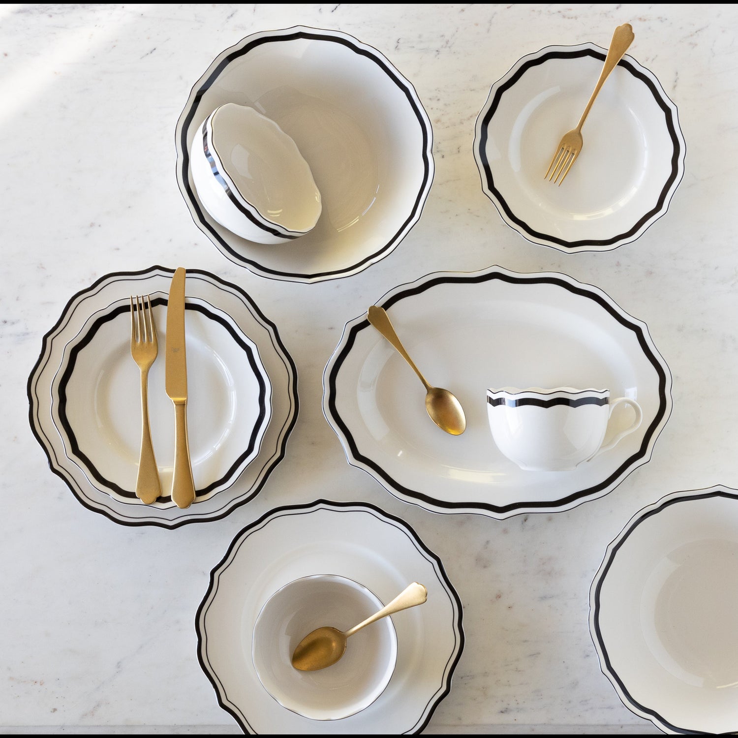Set of white ceramic dishes with black rims and gold cutlery on a light gray surface.