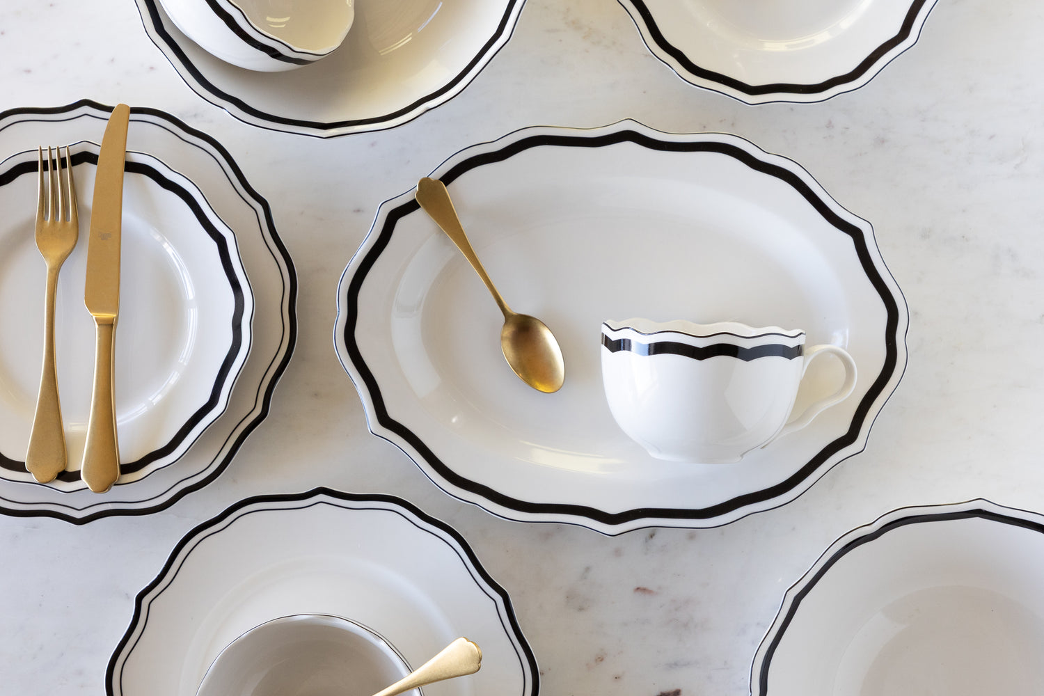 Set of white ceramic dishes with black rims and gold cutlery on a light gray surface.