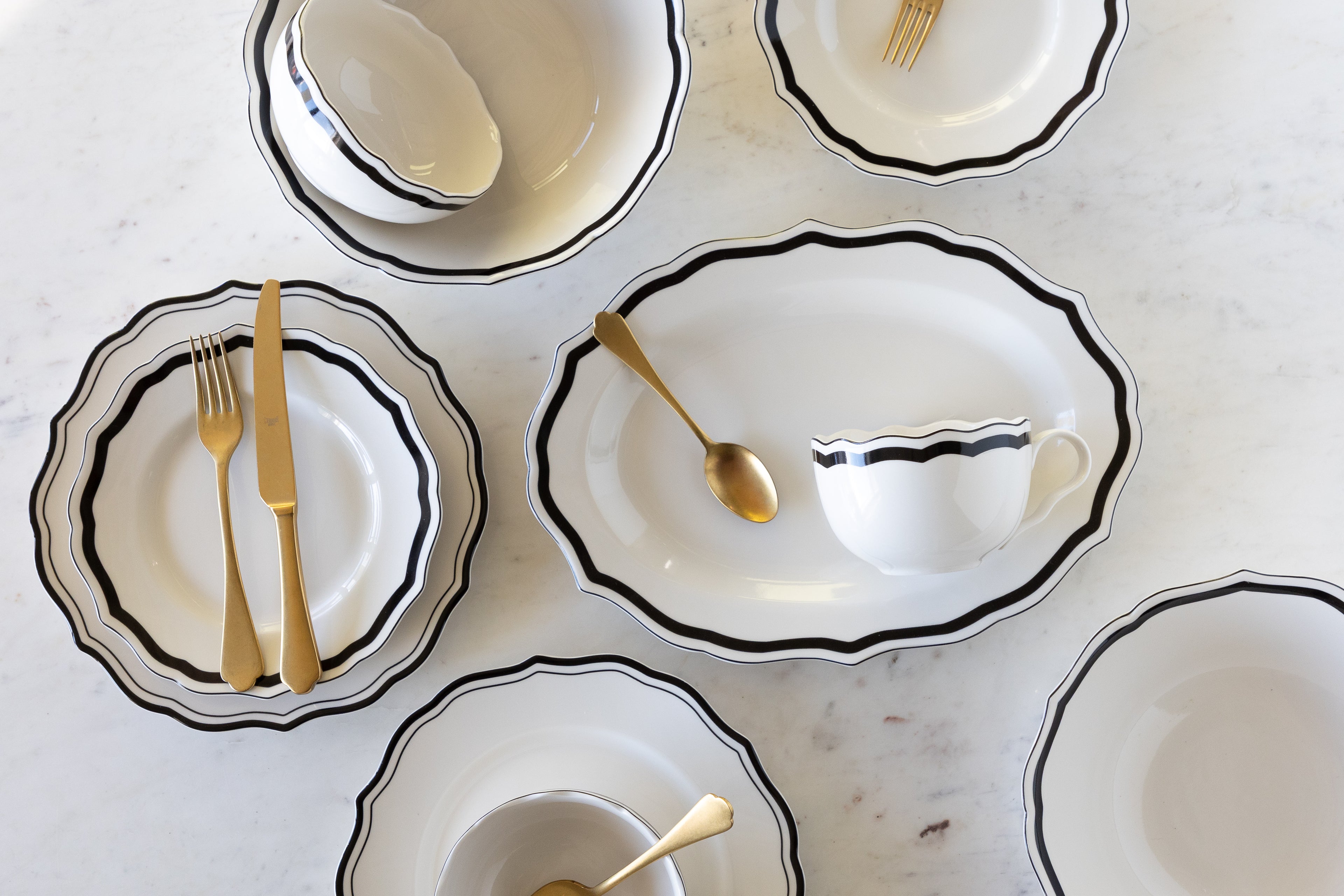 Set of white ceramic dishes with black rims and gold cutlery on a light gray surface.