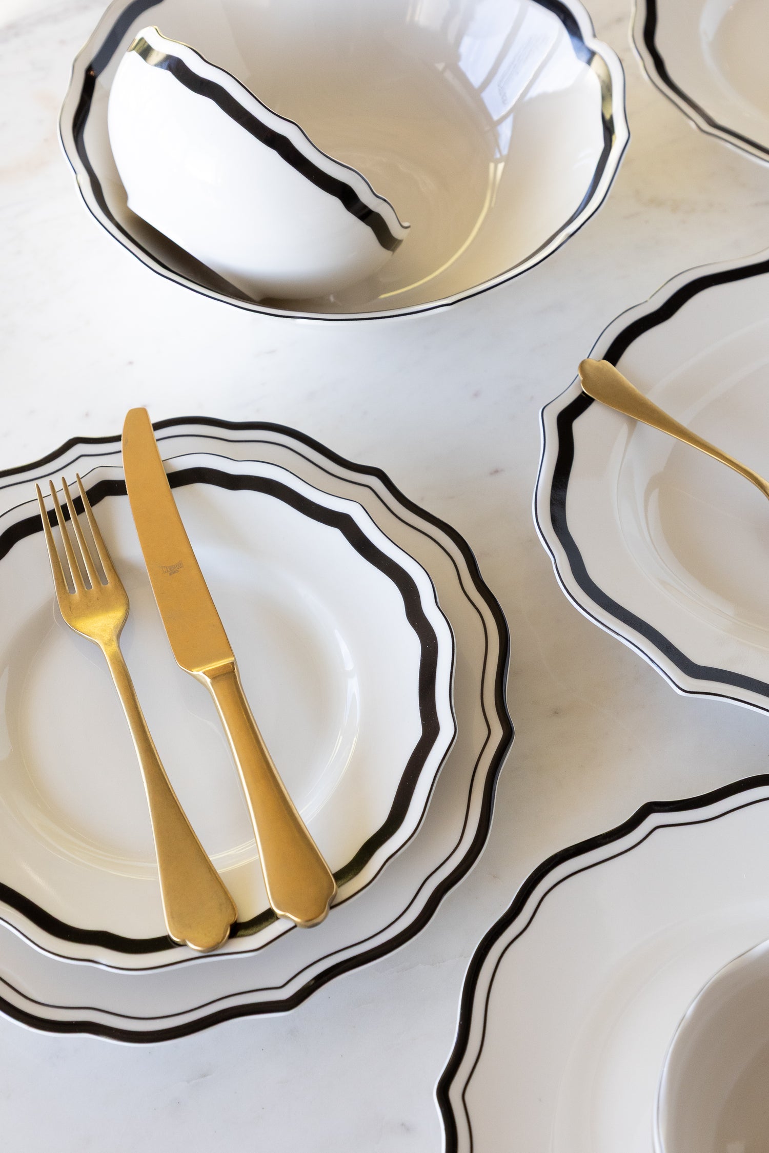 Set of white ceramic dishes with black rims and gold cutlery on a light gray surface.