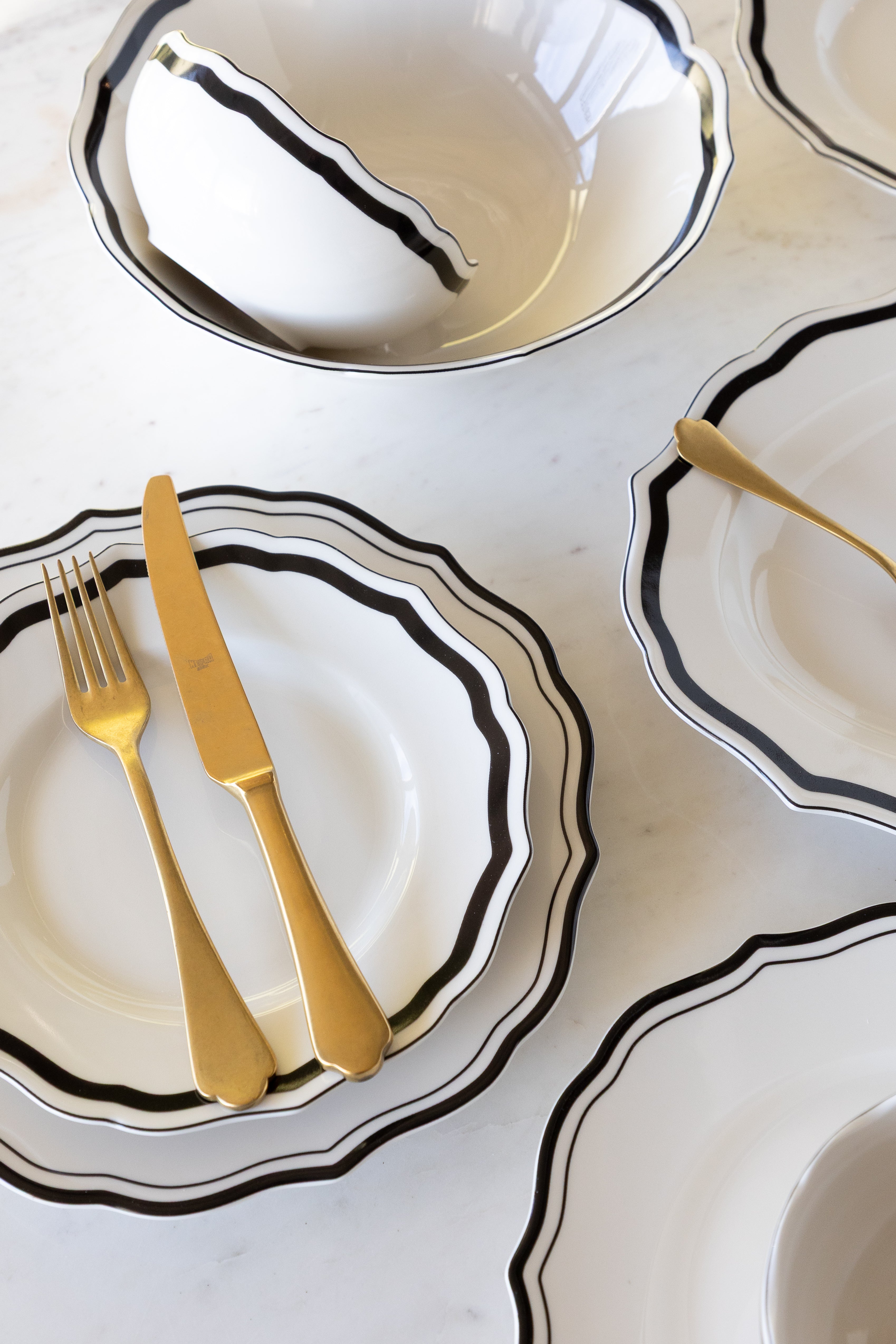 Set of white ceramic dishes with black rims and gold cutlery on a light gray surface.
