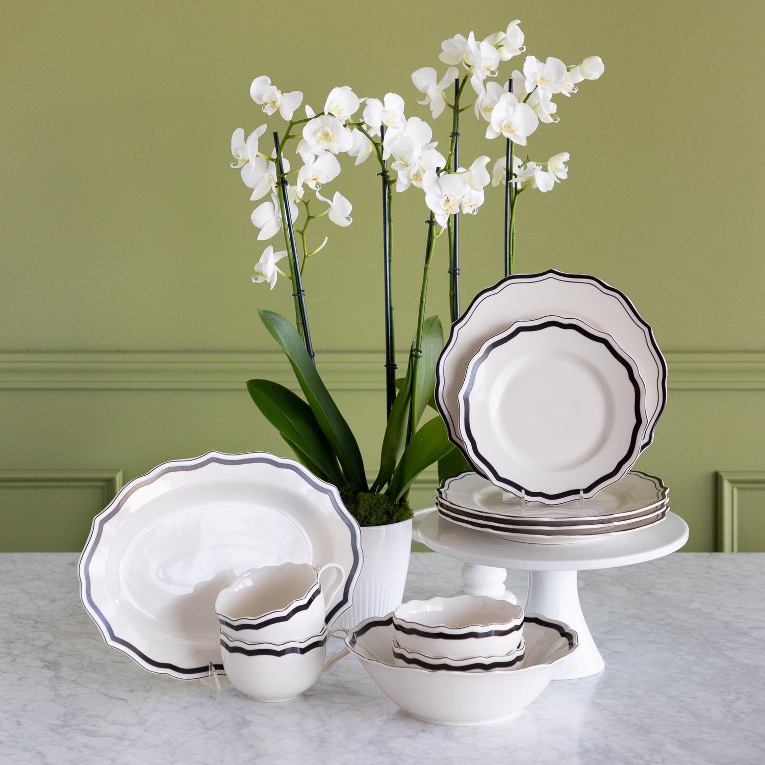 A white porcelain dinnerware set with black scalloped rims, including plates, bowls, cups, and a platter, arranged on a marble table with white orchids against a sage green paneled wall.