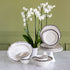 A white porcelain dinnerware set with black scalloped rims, including plates, bowls, cups, and a platter, arranged on a marble table with white orchids against a sage green paneled wall.
