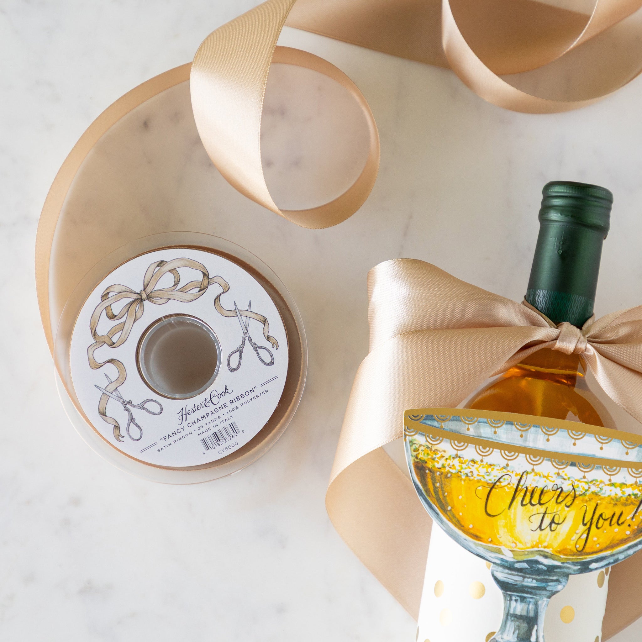 A bottle of wine with a gold bow, wrapped in the Gold Confetti Placemat and Champagne Coupe Place Card atop reading &quot;Cheers to You!&quot; sits beside a spool of Fancy Champagne Ribbon.