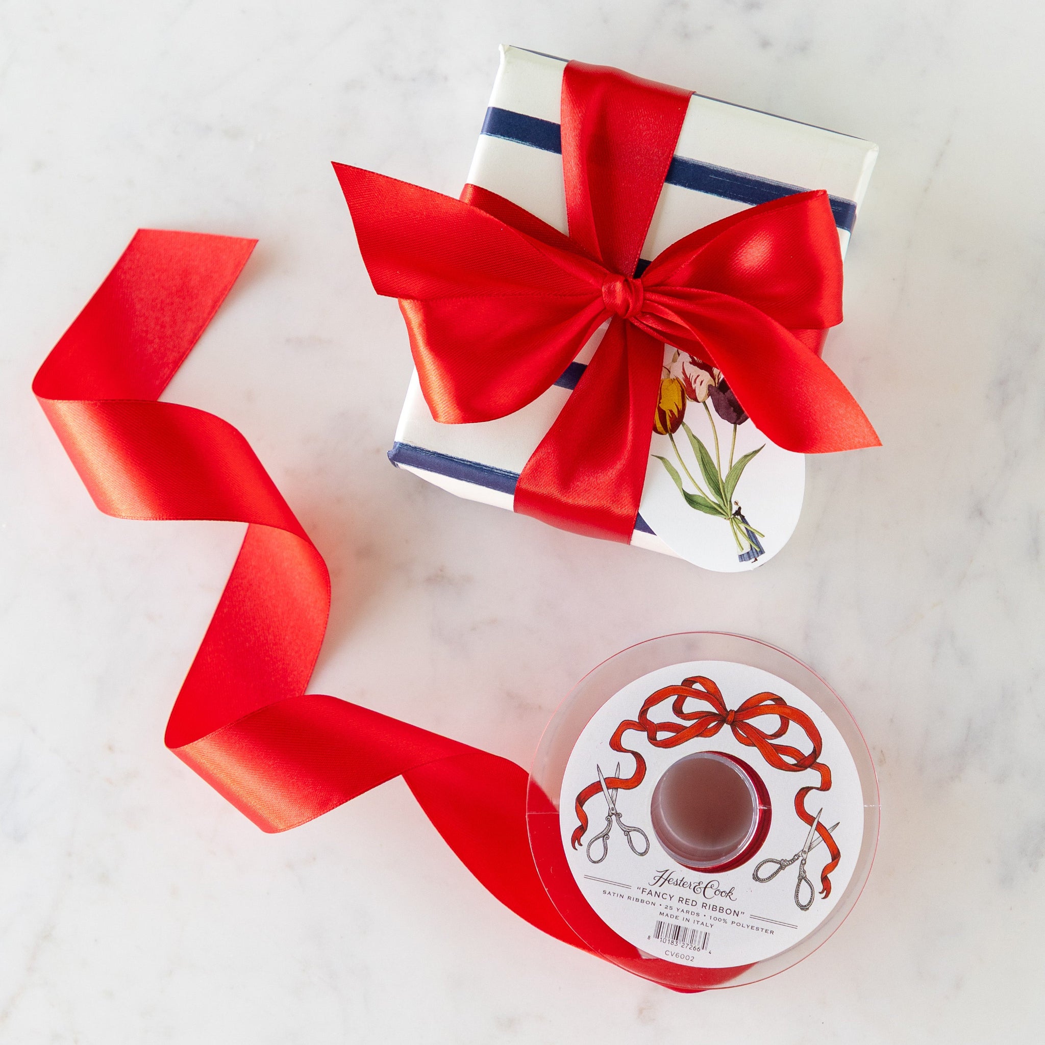 A present wrapped in a bow with the Fancy Red Ribbon featuring a Laura Stoddart gift tag, next to a spool of Fancy Red Ribbon.