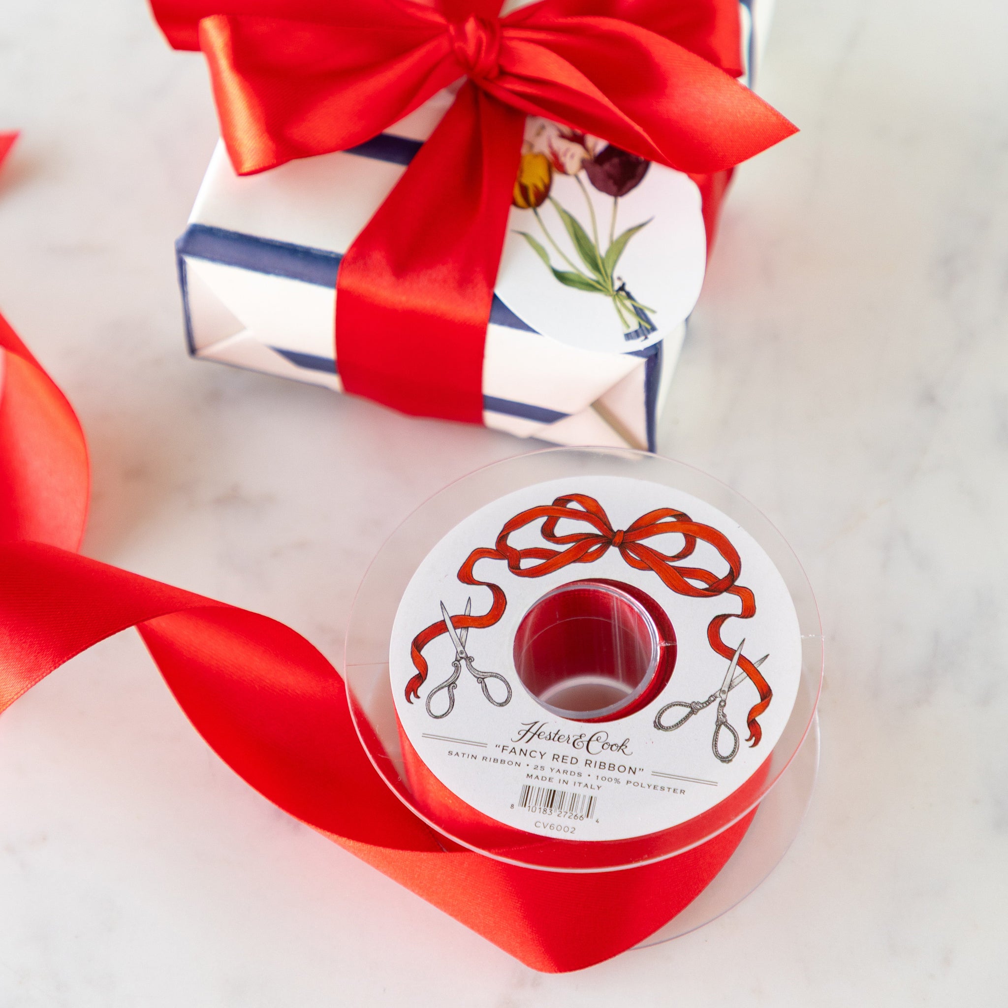 Spool of  Fancy Red Ribbon with a decorative label next to a wrapped gift with a red ribbon bow.