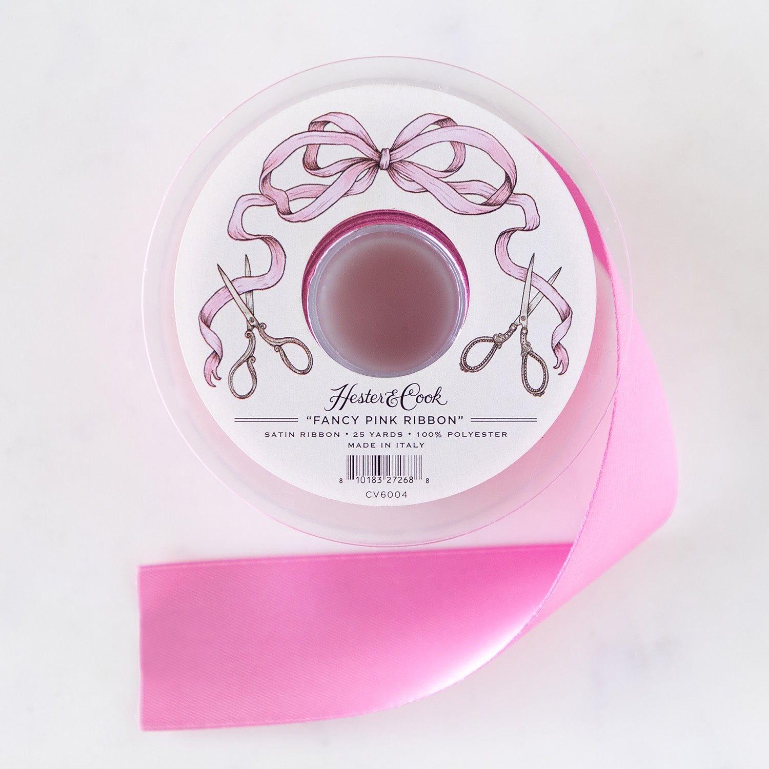 A spool of pink satin ribbon with a label featuring illustrated scissors and a pink bow, branded &quot;Hester &amp; Cook Fancy Pink Ribbon.