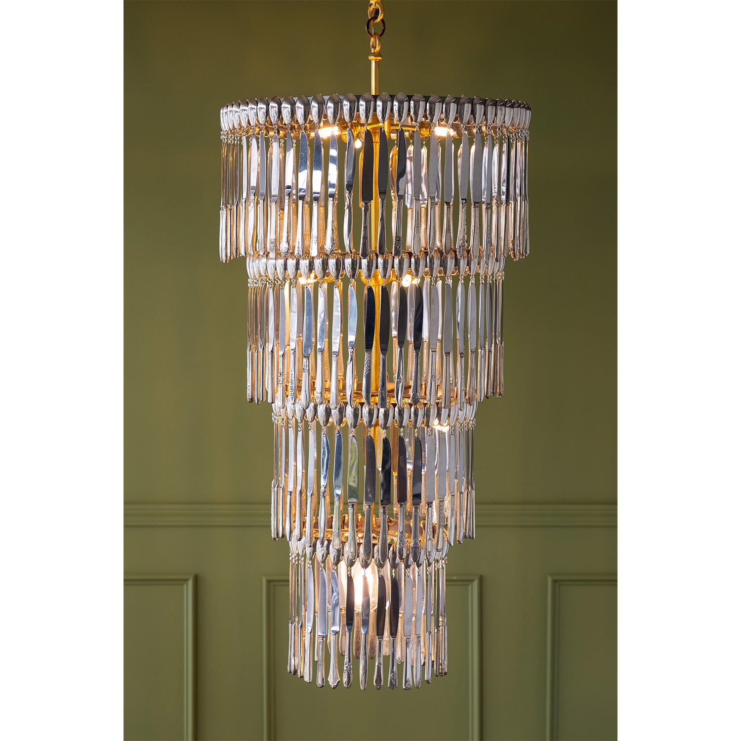 A tiered chandelier with vintage silver-plate knives and a gold-toned metal frame, hanging against an olive green paneled wall.