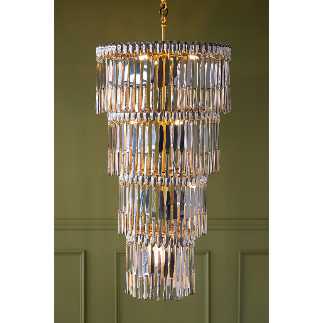 A tiered chandelier with vintage silver-plate knives and a gold-toned metal frame, hanging against an olive green paneled wall.