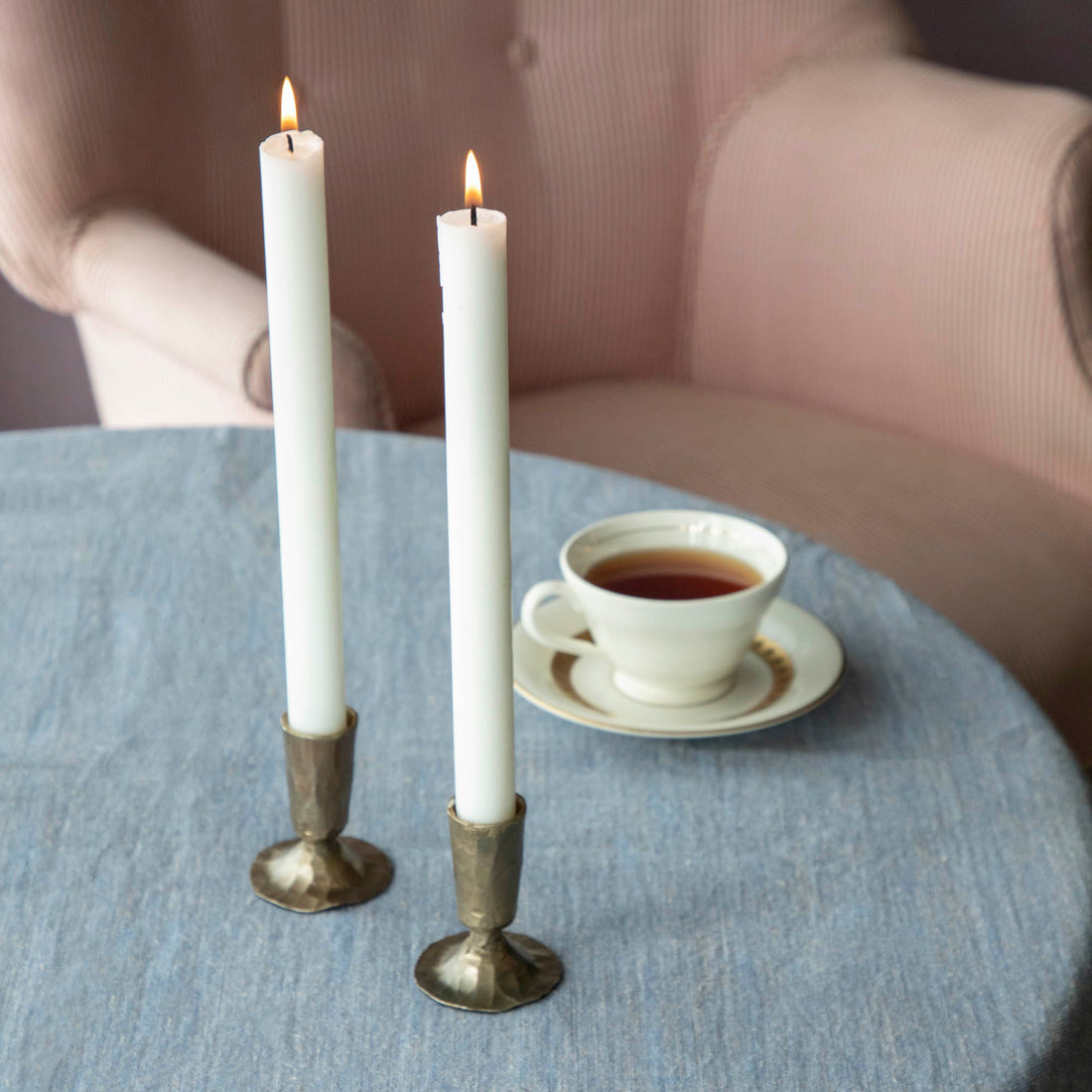 Two lit candles in Creative Co-Op metal taper holders on a table with a cup of tea in the background.