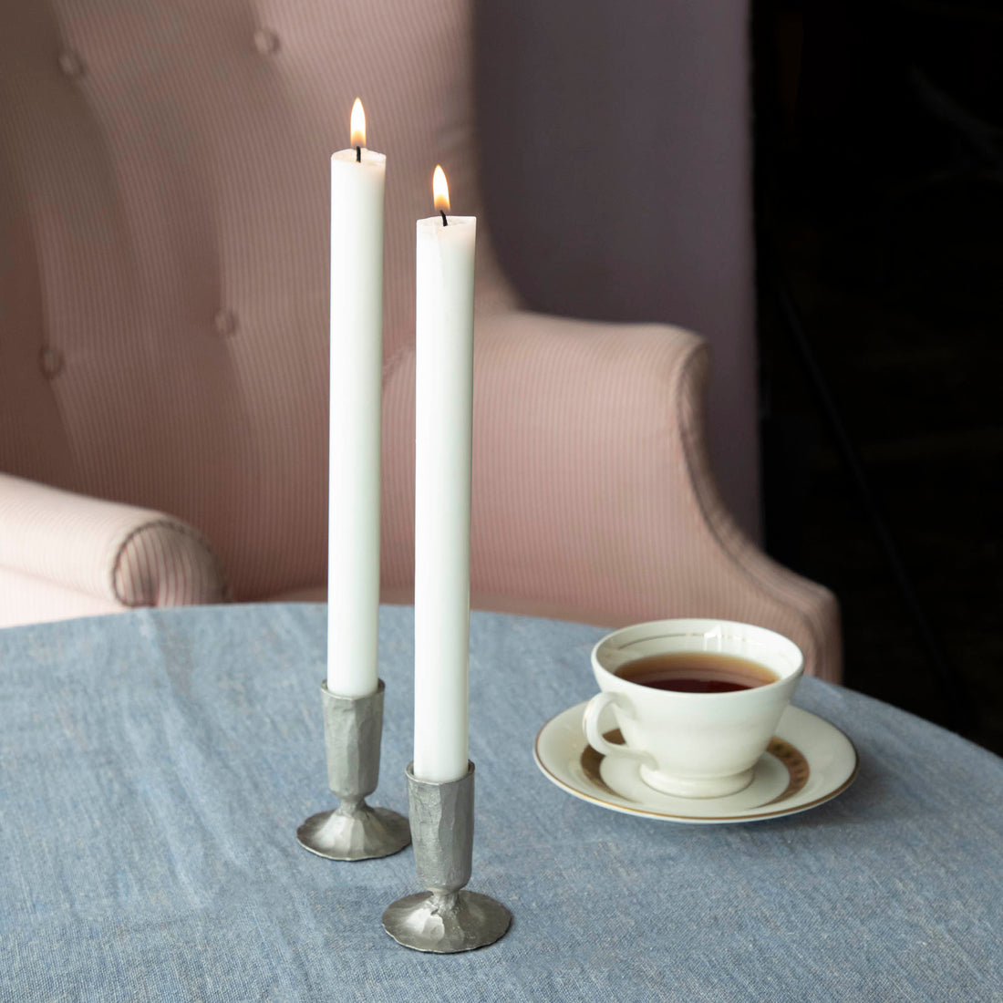 Two lit candles in Creative Co-Op metal taper holders on a table with a cup of tea in the background.