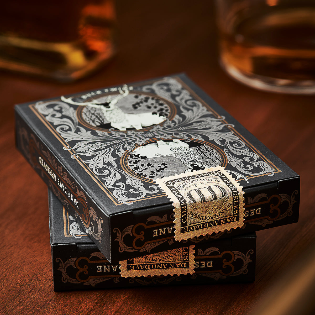 Decorative playing card boxes on a wooden surface with a glass of whiskey in the background.