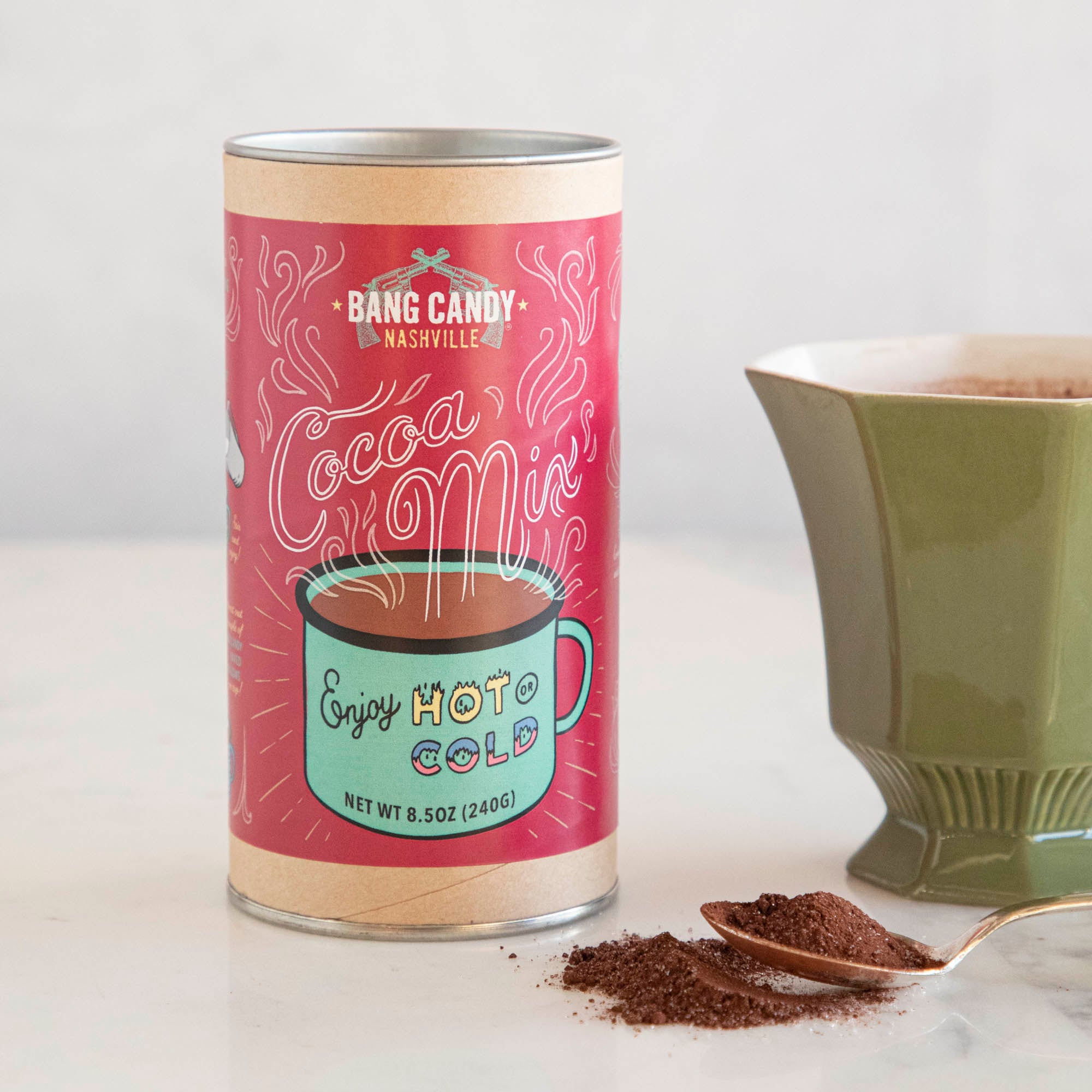 A tin of the popular Hester &amp; Cook Original Bang Candy Hot Cocoa Mix is positioned next to a steaming cup of coffee. The irresistible aroma and rich dark chocolate flavor of this hot cocoa mix make it the perfect