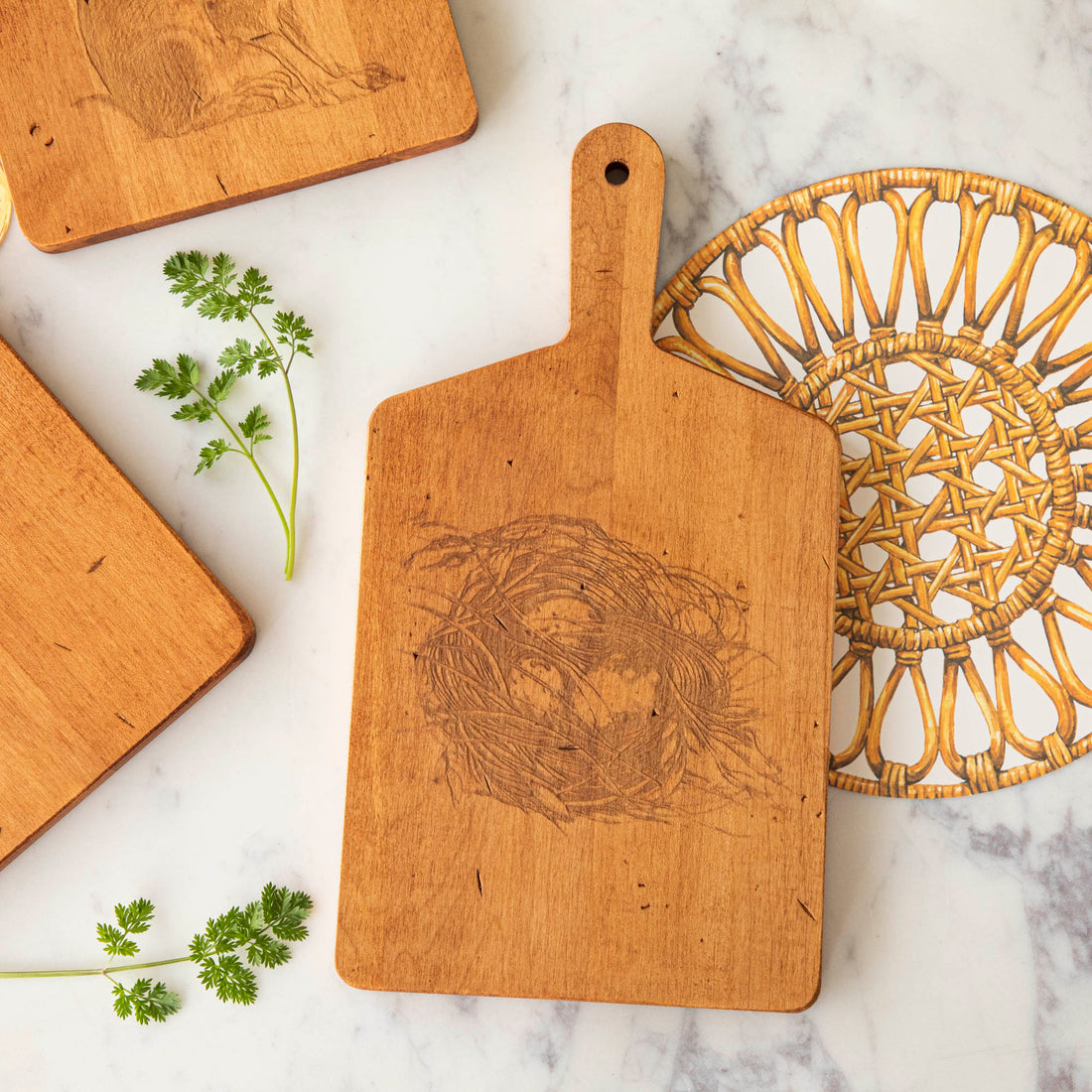 Wooden JK Adams Nest Artisan Maple Rectangle Cheese Board with visible knife marks next to a woven trivet and fresh herbs on a marble countertop.