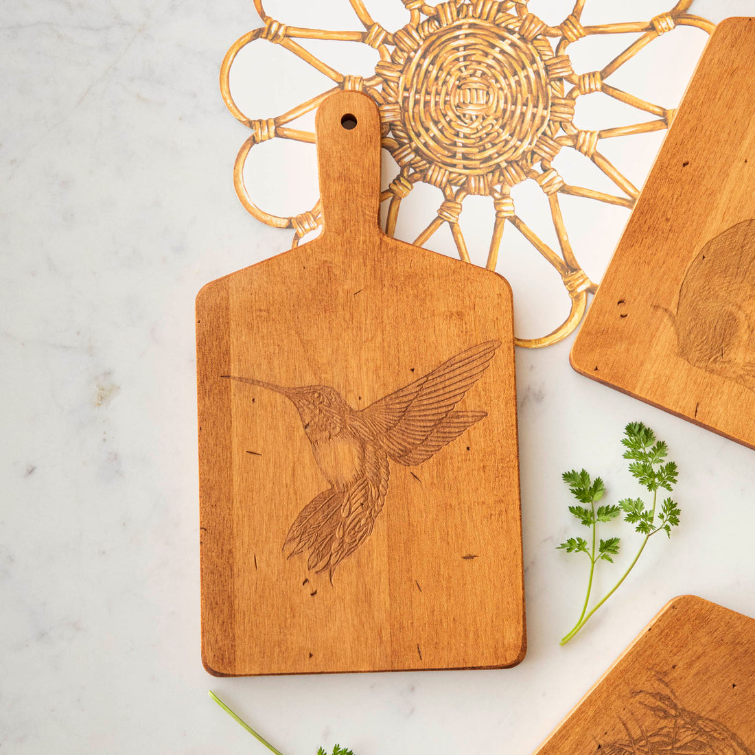 Artisanal Hummingbird Artisan Maple Rectangle Cheese Board with an etched hummingbird design on a marble countertop beside decorative items and herbs by JK Adams.