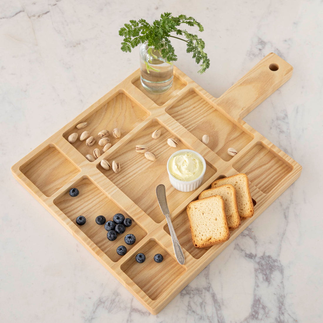 A JK Adams Ash Divided Serving Board with compartments holding pistachios, blueberries, crackers, and a dip, next to a small vase with greenery.